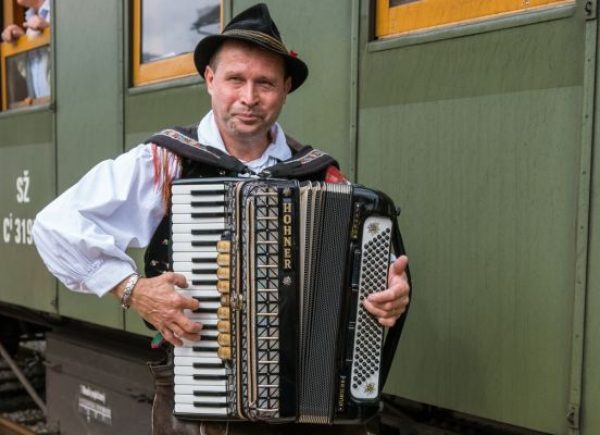 Man dressed in the Gorenjska national costume playing accordion on the Jesenice train station in front of the museum train car